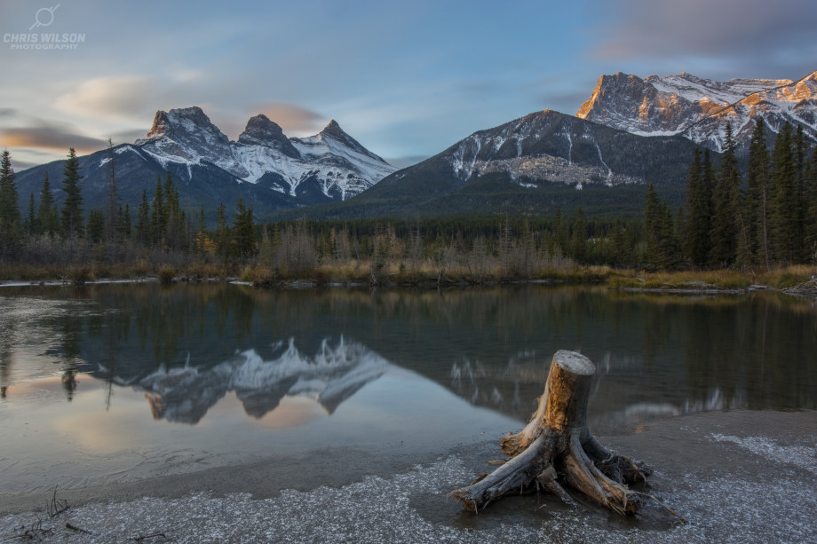 Tall snowy and rocky mountains glow with sunset light above forests of pine, all reflected in a still lake in the foreground.