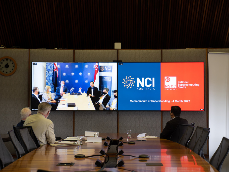 Long view along a boardroom table. Three men are seated at the end of the table looking at television screens showing the NCI and NSCC logos, and the other attendees of the video conference they are participating in.