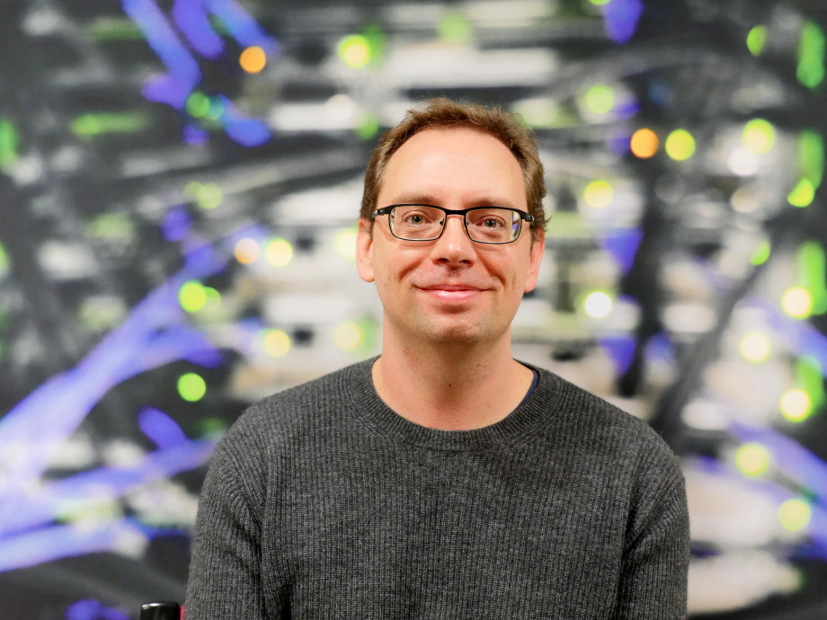 A man in a dark t-shirt and black-rimmed glasses in front of a blurred background of computer cables and lights.