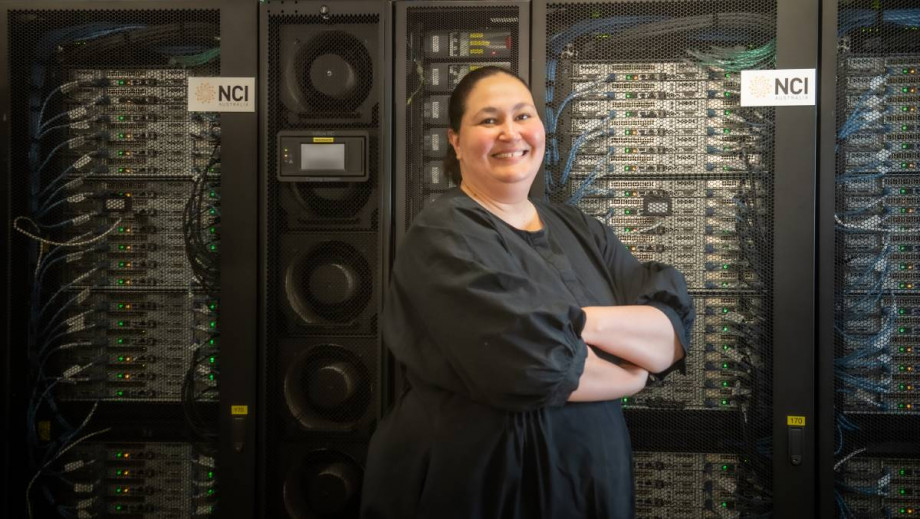 A woman in a black dress, Azure Hermes, stands with her arms crossed smiling at the camera. She is standing in front of a wall of computer servers with flashing lights and cables, with the NCI Australia logo visible.