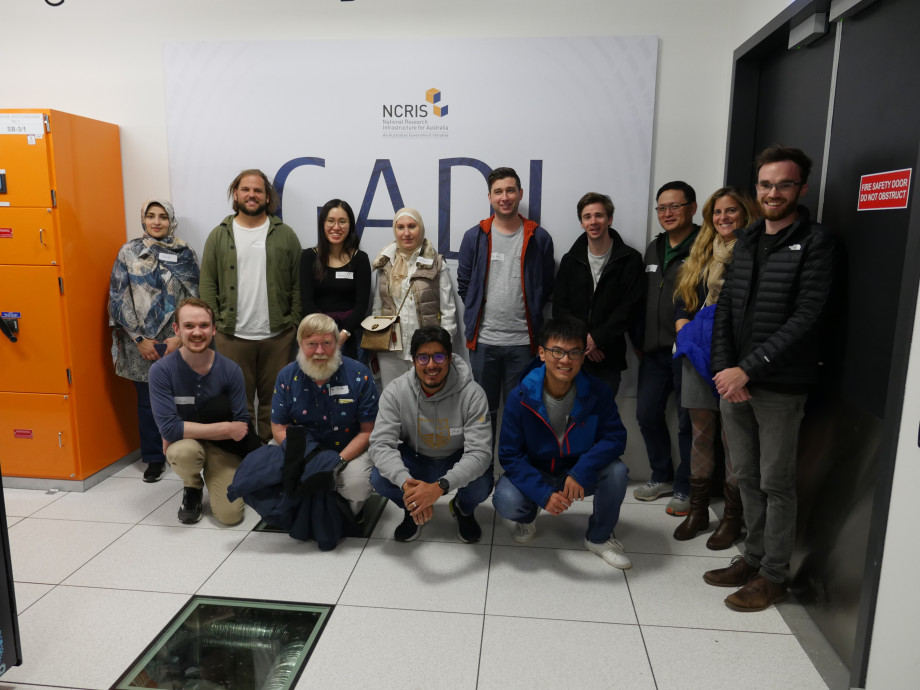 A group of a dozen people standing and crouching in front of a big sign on a wall reading GADI with some letters partially obscured.