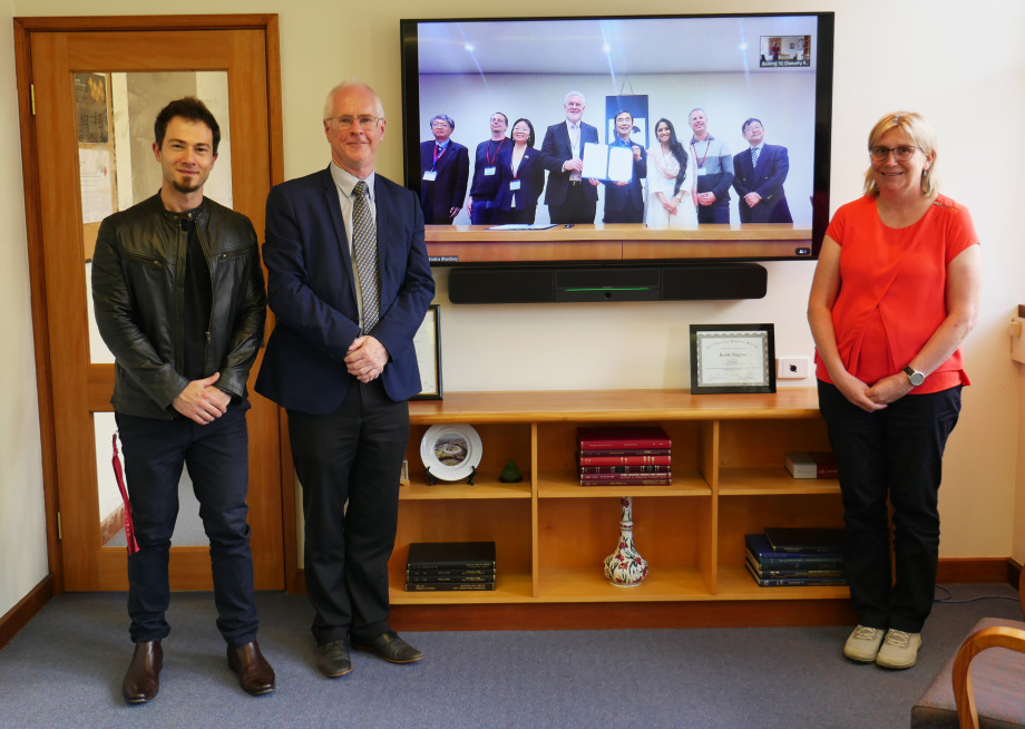 Three people standing on either side of a big television on a wall. The television shows eight people standing together, with two in the middle holding some documents. All the people are smiling for the camera.