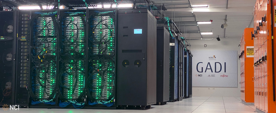 Long view of a data centre, with multiple pods of server racks all visible in a row. Bright green lights are visible on the nearest pod, and against the back wall is a sign that says Gadi in big letters.