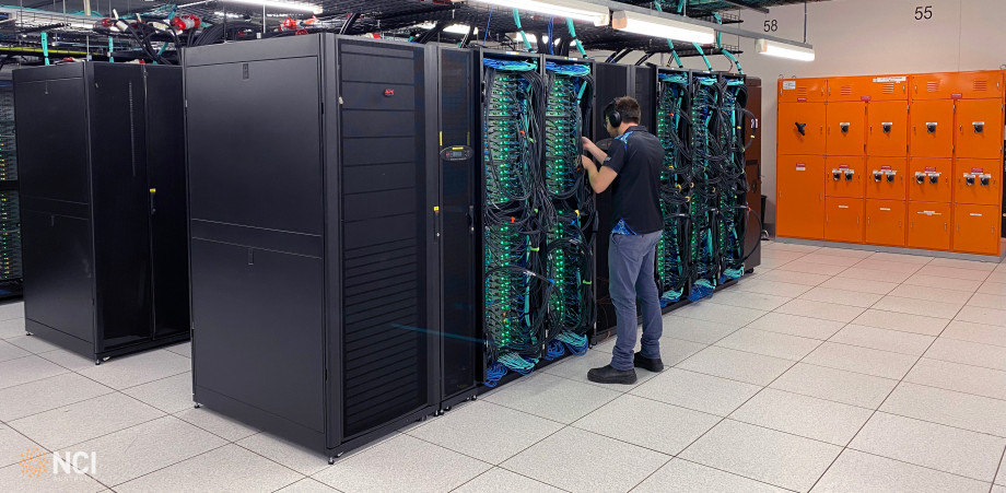 A technician working on the cabling in a pod of computer servers with green lights and complex cabling.