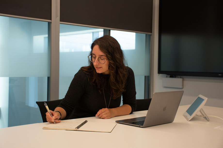 A woman with curly brown hair sits at a laptop, with a notepad next to the computer which is writing in.