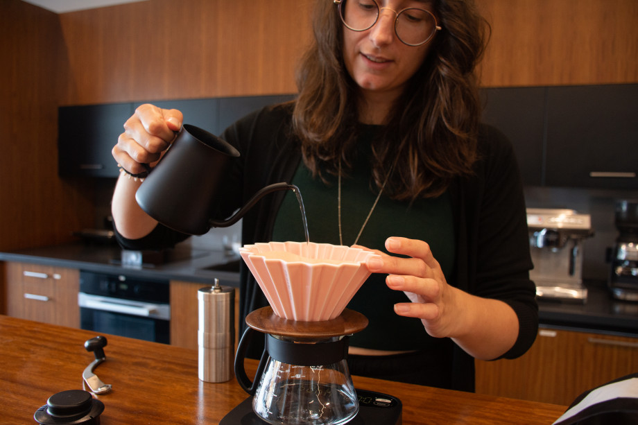 A woman with curly brown hair is pouring water into a folded paper funnel sitting over a mug as she brews some coffee.