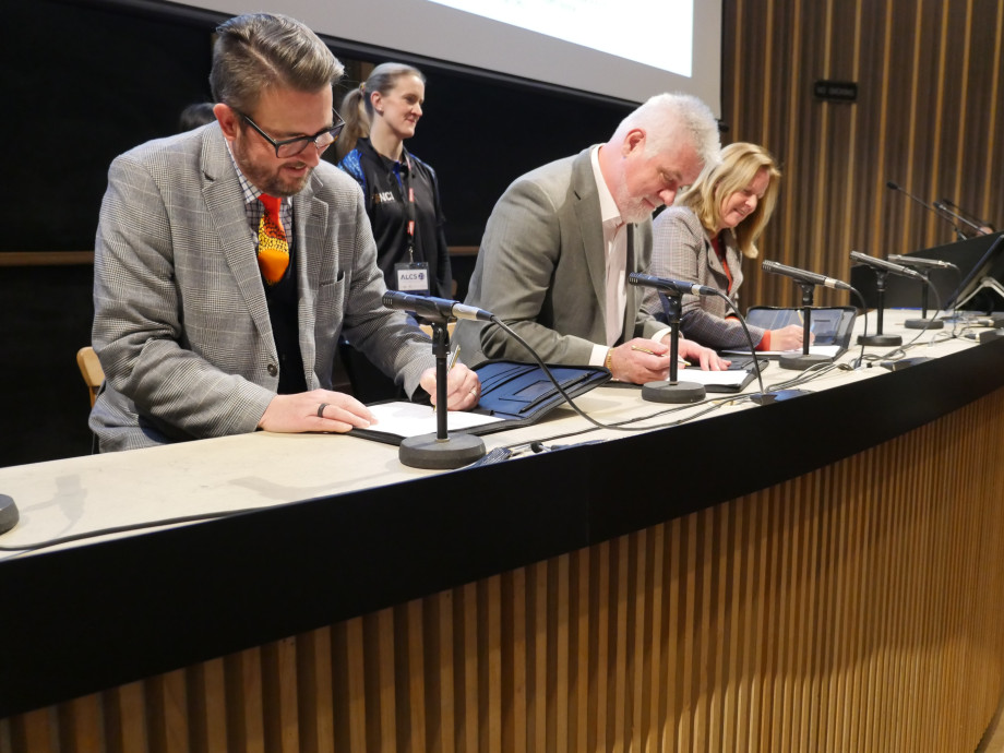 Three people at a long desk in the front of an auditorium signing documents while smiling.