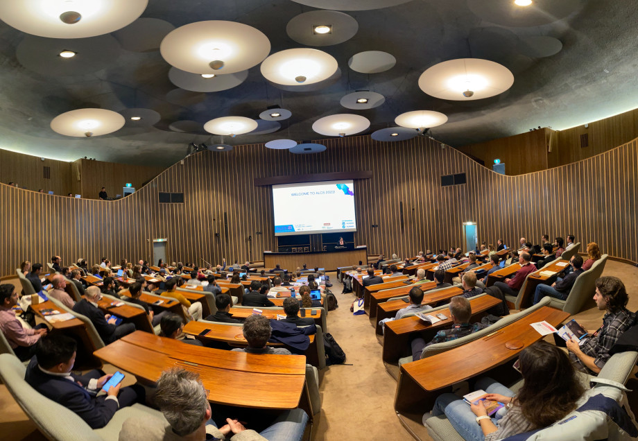Wide view of a full, beautiful auditorium with wooden desks and carpeted floor, with a woman speaking under a big projector screen saying "Welcome to ALCS2023".