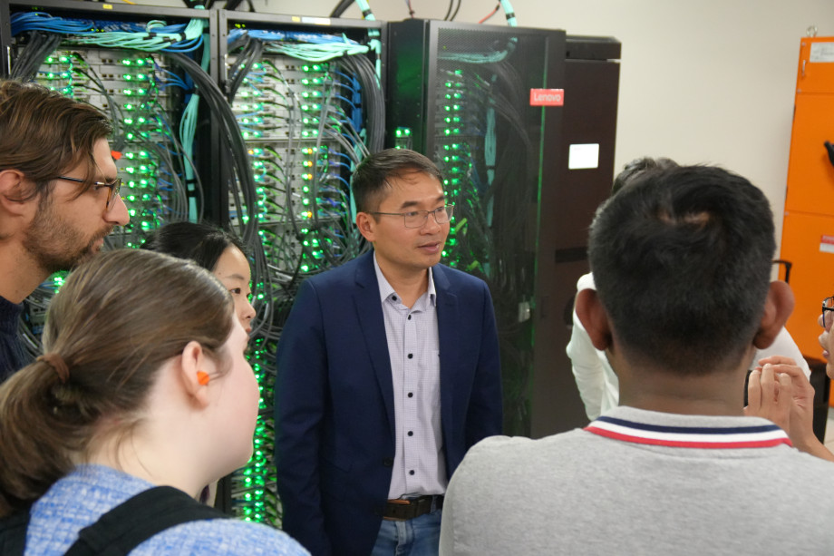 A man in a blue suit stands in front of a supercomputer with green lights. Facing him there are a number of people listening to him whose backs we can see.