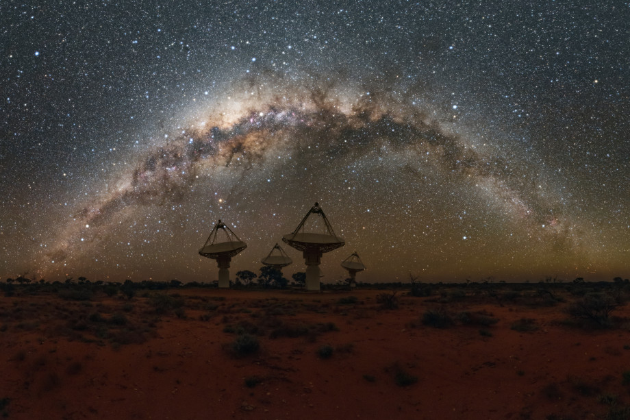 Desert landscape at night, with four radio satellite dishes visible on the horizon and many stars in the sky.