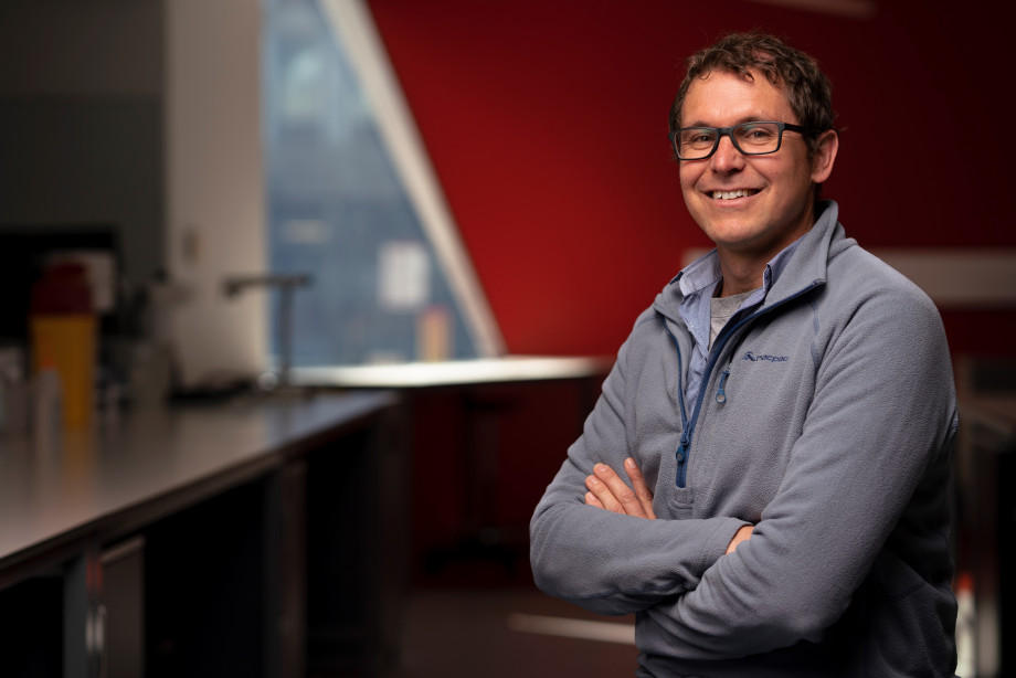 A man, Ben Corry wearing glasses and a jumper stands in a science laboratory with his arms crossed smiling at the camera.