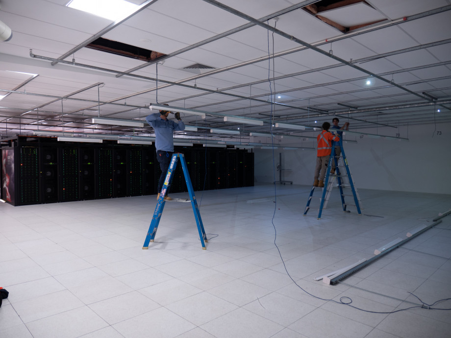 Men on ladders removing long fluorescent lights hanging from the ceiling a large room with a supercomputer in the background.