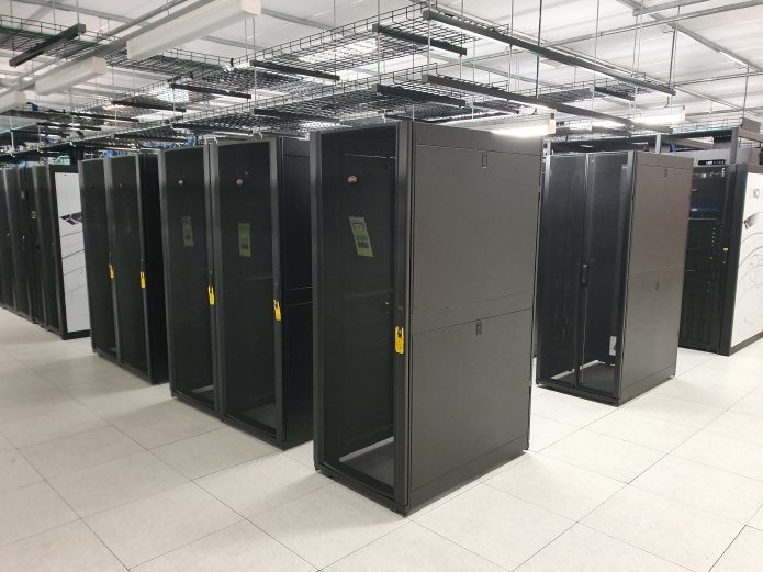 Six black server racks stand empty in rows surrounded by functioning supercomputer equipment. 