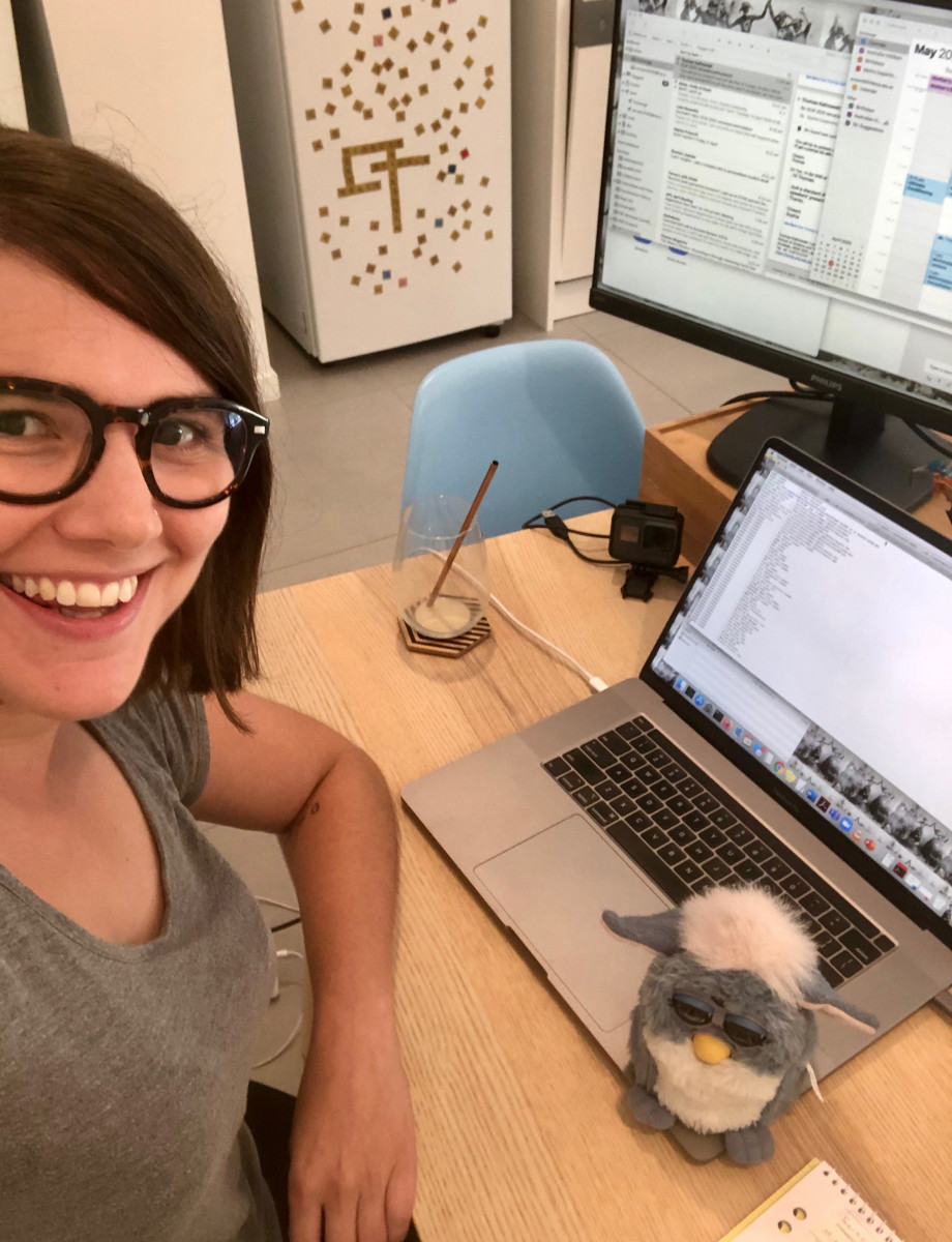 Sophie sits at a table smiling up at the camera. On the table are a laptop, monitor and a furby. In the background is a fridge.