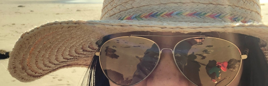 Close up of Jin wearing sunglasses and a hat at the beach. In the reflection of the sunglasses is Jin's daughter playing in the sand.
