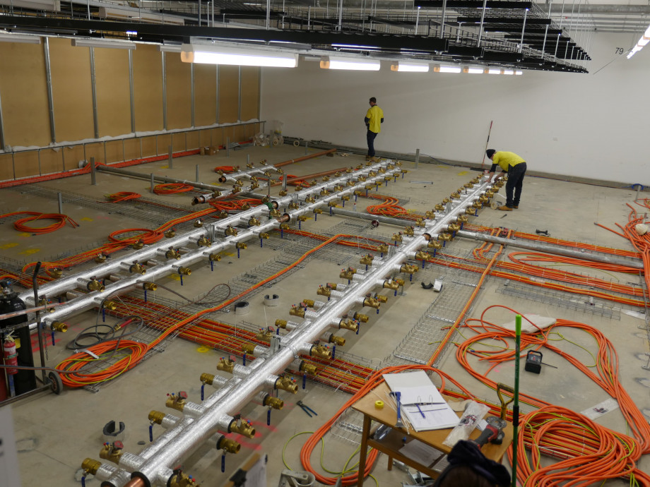 A concrete floor with neatly arranged orange electrical cables, now with long lines of metal pipes in place along the floor.