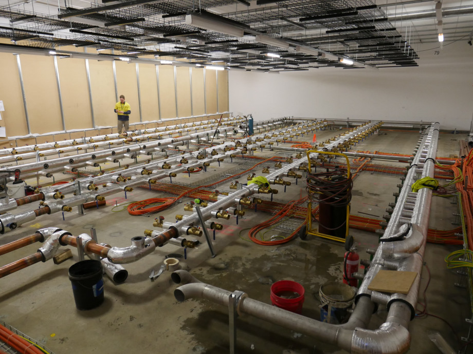 A concrete floor with orange electrical cables arranged in a neat grid, now with a full complement of silver metal pipes arranged in rows above the cables.