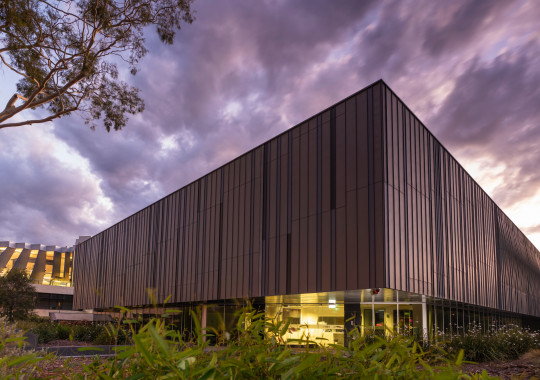 A rectangular building with striking vertical stripes in shades of grey and brown viewed from an angle, with hazy purple clouds above.