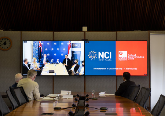 Long view along a boardroom table. Three men are seated at the end of the table looking at television screens showing the NCI and NSCC logos, and the other attendees of the video conference they are participating in.