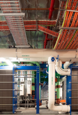 Blue and grey machines in front of white, green and blue pipes, and neatly arranged lengths of orange electrical cables.