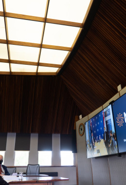 A wide view of a room with three men at the end of a boardroom table looking at television screens on which the NCI and NSCC logos are displayed, and with other participants of the signing ceremony also visible.