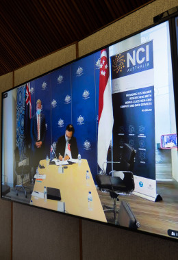 A television screen with two men visible, one standing in front of Australian and Singaporean flags, and the other sitting at a desk signing an important document.