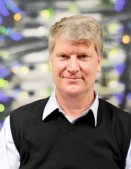 Ben Evans smiling facing the camera in front of a blurry background of computer lights and cables.