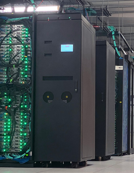 Long view of a data centre, with multiple pods of server racks all visible in a row. Bright green lights are visible on the nearest pod, and against the back wall is a sign that says Gadi in big letters.