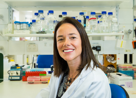 Carola Vinuesa sits smiling at the camera in front of a lab bench filled with jars and other scientific equipment.