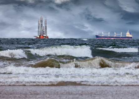An oil rig and shipping container off shore from a beach while dark storm clouds approach.