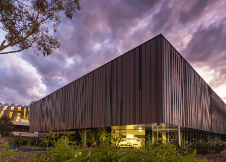 A rectangular building with striking vertical stripes in shades of grey and brown viewed from an angle, with hazy purple clouds above.