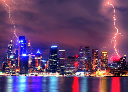 Two bolts of lightning strike a major city at night time. Lights of tall buildings are all visible and the whole image has a purple tint.