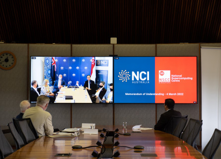 Long view along a boardroom table. Three men are seated at the end of the table looking at television screens showing the NCI and NSCC logos, and the other attendees of the video conference they are participating in.