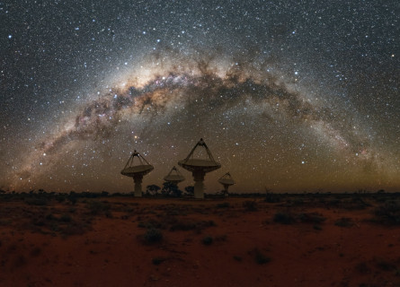 Desert landscape at night, with four radio telescope dishes visible on the horizon and many stars in the sky.  