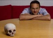 A human skull on a wooden table filling the frame, with a man behind the table leaning on it with arms crossed.