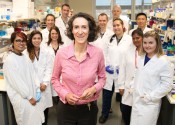 Sally Dunwoodie stands smiling at the camera surrounded by colleagues in white lab coats.