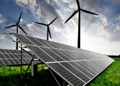 Solar panels on green grass in front of three wind turbines silhouetted by the sun behind them.