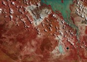 Screenshot from the Open Air video art project by Grayson Cooke. The video shows time lapse image sequences of the Australian landscape made using satellite imagery, interspersed with macro photography of paintings being created. The screenshot shows a river and lake in false colour blue, with lots of tiny clouds and the red outback.