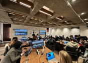 A seminar room filled with around 8 tables at which 3 or 4 people are all sitting. There is a presenter at the front of the room showing computer code on a big screen. 