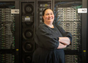 A woman in a black dress, Azure Hermes, stands with her arms crossed smiling at the camera. She is standing in front of a wall of computer servers with flashing lights and cables, with the NCI Australia logo visible.