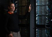A woman stands in a dark space with her hand on the door of a supercomputer cabinet. She and the complex cables in the supercomputer are brightly lit.