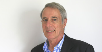 Greg Ayers, wearing a dark suit and blue and white shirt, smiles at the camera.