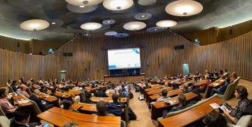 Wide view of a full, beautiful auditorium with wooden desks and carpeted floor, with a woman speaking under a big projector screen saying "Welcome to ALCS2023".