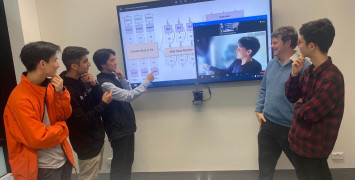 Group photo of men standing in front of a large television pointing to one remotely connected colleague on the screen. Everyone is posing for a photo.