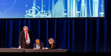 Three people sitting at a desk on stage. The middle one is signing a piece of paper.