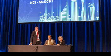 Three people sitting at a desk on stage. The middle one is signing a piece of paper.